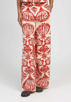 Pantalon à jambes larges avec motifs floraux complexes rouges et crème, porté avec un haut assorti et des sandales noires tissées.