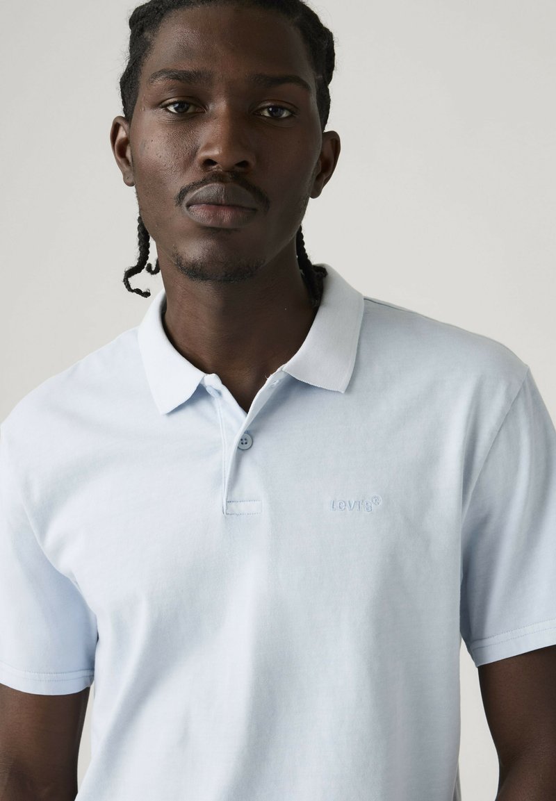 Hombre con cabello trenzado que lleva una camiseta polo azul claro de Levi's, mirando hacia adelante sobre un fondo claro y sencillo.