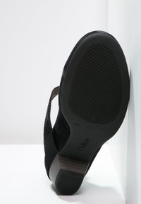 Chaussure en daim noir avec un petit talon carré, semelle texturée et un design découpé révélant la doublure intérieure. Finition lisse et mate.