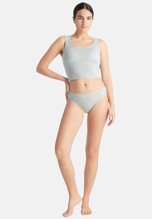 Frau steht barfuß, trägt ein hellgraues Crop-Top und passende Unterwäsche, posiert mit einer Hand in der Hüfte vor weißem Hintergrund.