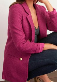 Blazer texturé fuchsia avec revers à encoches, boutons dorés et détail de manchette intérieure rose, associé à un jean foncé et un top en dentelle.