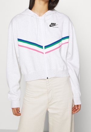 Biała bluza Nike zapinana na zamek z niebieskimi, zielonymi, białymi i różowymi paskami w jodełkę, noszona z kremowymi spodniami na stojącej osobie.