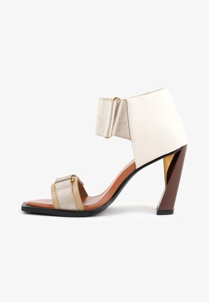 Sandalia de tacón alto con una correa de cuero beige y herrajes dorados. Presenta una amplia correa en el tobillo, un tacón triangular y un panel lateral texturizado.