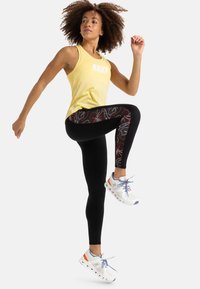 Gelbes Tanktop mit "ROXY"-Druck, schwarze Leggings mit buntem Blumenmuster und weiße Sportschuhe mit orangefarbenen und blauen Akzenten.