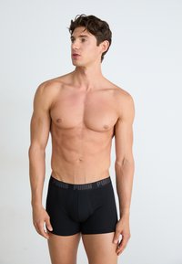 Boxeri negri dintr-un material moale, cu un tiv elastic lat pe talie, având un logo repetat. Textură netedă și design mulat.