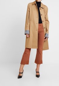 Manteau trench beige avec des poignets à carreaux noir et blanc, associé à un pantalon évasé couleur rouille avec un ourlet festonné. Des talons noirs complètent la tenue.