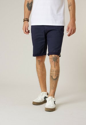Man draagt marineblauwe, gestructureerde korte broek, witte sneakers en een wit overhemd, met zichtbare tatoeages op armen en benen.