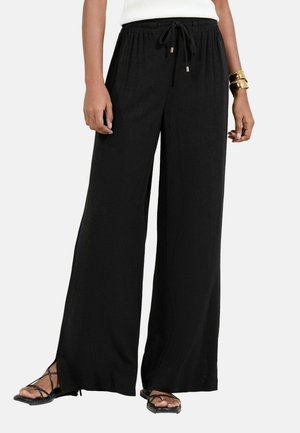 Moteris dėvi juodas plačias kelnes su raišteliu liemens srityje, derina jas su juodais sandalais su dirželiais ir auksiniais apyrankėmis dešinėje riešo pusėje.