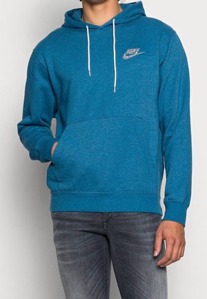 Mann trägt blauen Nike-Hoodie mit weißen Kordeln und Fronttasche, kombiniert mit grauen, ausgewaschenen Jeans, eine Hand in der Tasche.