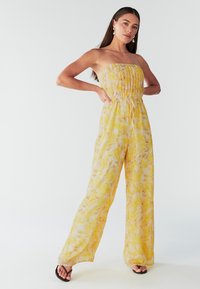 Une combinaison sans bretelles en tissu léger et fluide, de couleur jaune avec un motif floral, présentant un corsage smocké et un design à jambes larges.