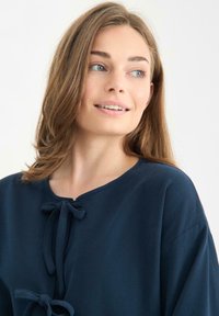 Granatowa koszula z okrągłym dekoltem i dwoma kokardkami z przodu. Miękka tkanina o gładkiej fakturze i luźnym kroju.