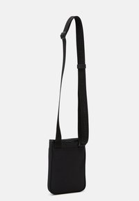 Sort nylon crossbody taske med flad design, justerbar rem og struktureret kant. Har logo detaljer på remmen.