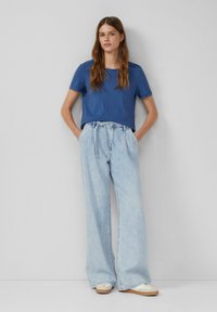 T-shirt en coton bleu à manches courtes associé à un pantalon en denim large à taille haute avec un lien. Le mannequin se tient avec les mains dans les poches.