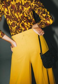 Pantaloni gialli a gamba larga con vita alta e tasche posteriori, abbinati a un top a maniche lunghe marrone e giallo con fantasia e una piccola borsa nera.