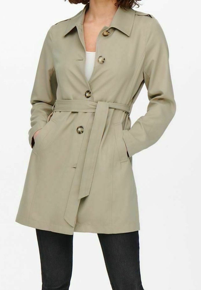 Femme portant un trench-coat beige ceinturé avec des boutons, mains dans les poches, assorti à un pantalon noir et un haut blanc en dessous.
