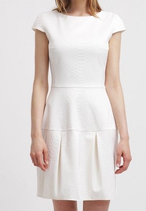 Frau trägt ein weißes, strukturiertes Kurzarmkleid mit tailliertem Schnitt und plissiertem Rock, stehend vor einem schlichten Hintergrund.