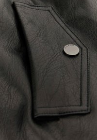Zwart faux leren stof met een gestructureerd oppervlak, voorzien van een zilveren drukknoop met "GUESS" gegraveerd, en een gestikt zakcontour.