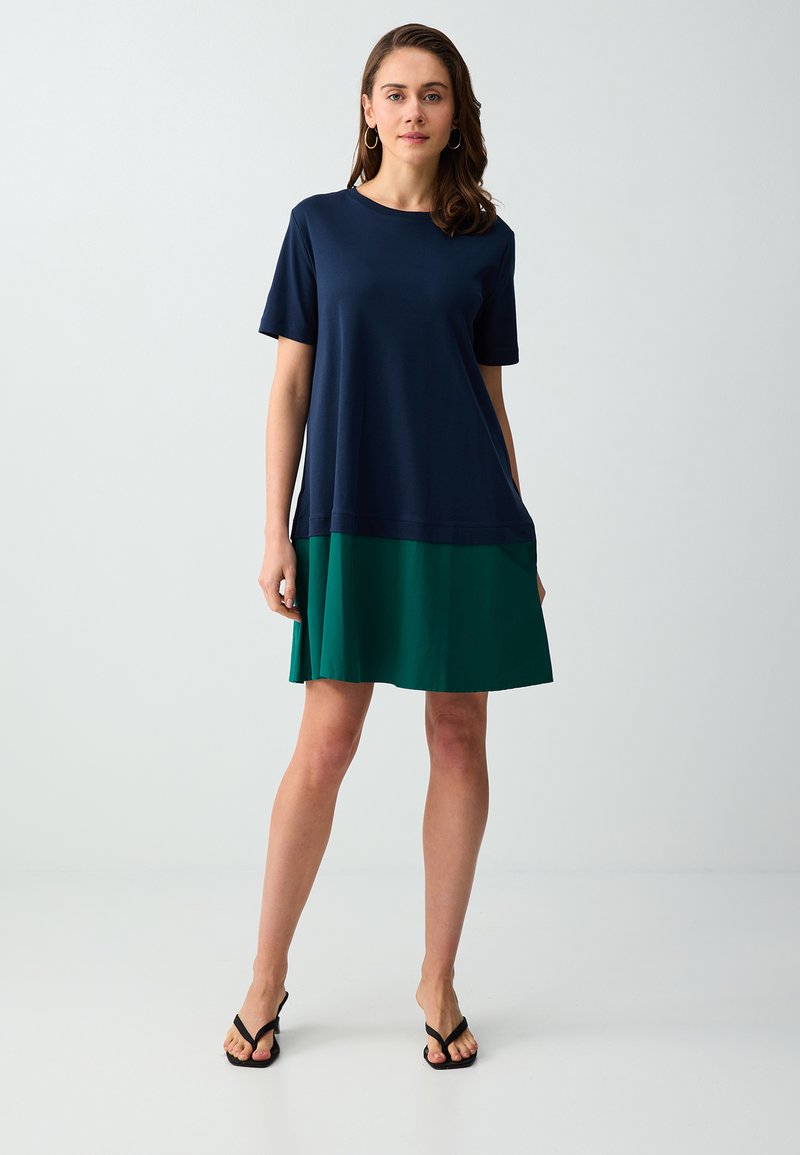 Femme portant une robe courte bleu marine et verte à manches courtes, avec des talons ouverts noirs, se tenant devant un fond gris clair uni.