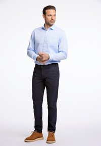 Lichtblauw overhemden met borstzak, gedragen met donkerblauwe chino's en bruine suède schoenen met witte zolen. Eenvoudige achtergrond.