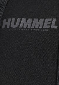 Schwarzer Sportbekleidungsstoff mit dem "HUMMEL"-Logo in einem etwas helleren Farbton, mit glatter Textur und subtiler Strickmusterung.