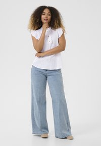 Weiße Leinenbluse mit kurzen, fransigen Ärmeln und Knopfleiste. Kombiniert mit hellblauen, weit geschnittenen Jeans und beigen Schuhen.