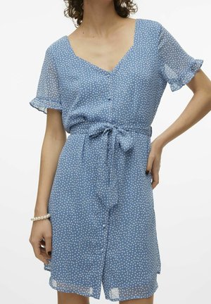Kvinde iført blå polka dot kortærmet kjole med knapper og bundet bælte i taljen, den ene hånd på hoften, den anden synlig med perlearmbånd.