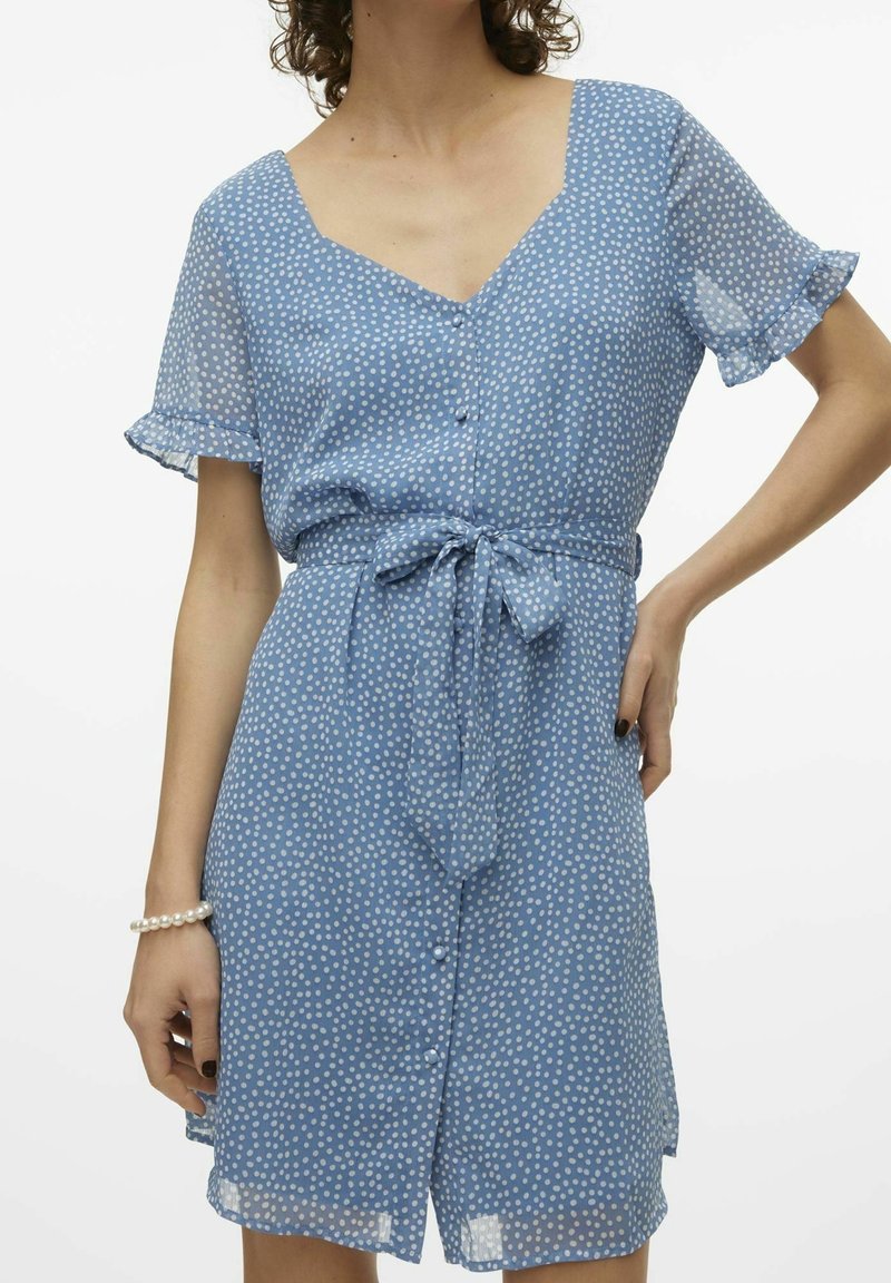 Femme portant une robe bleue à pois, manches courtes, avec des boutons et une ceinture nouée à la taille, une main sur la hanche, l'autre visible avec un bracelet de perles.