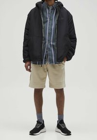 Czarna kurtka puchowa noszona na niebieskiej i zielonej koszuli w kratę, beżowych szortach i czarnych trampkach z białymi podeszwami oraz szarymi skarpetami.