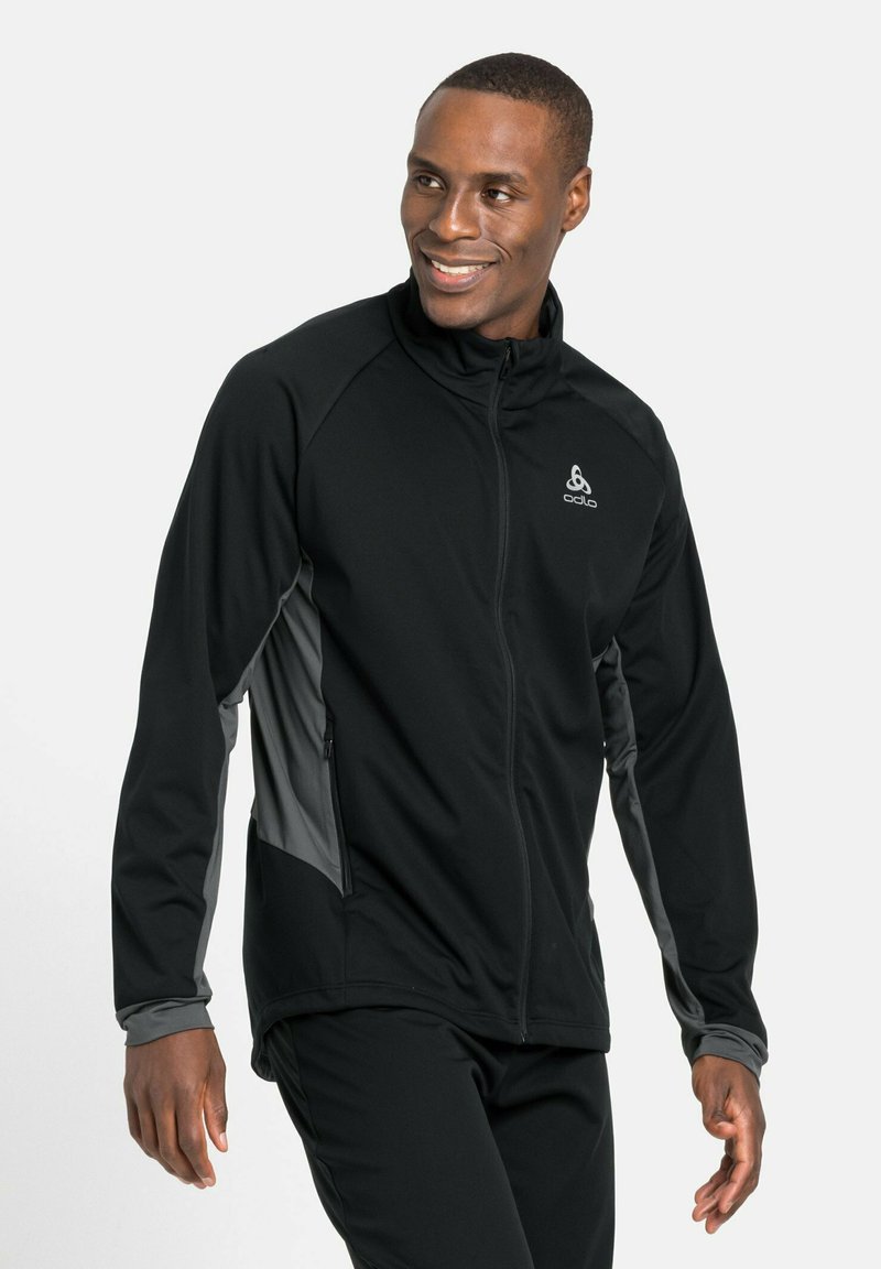 Schwarze Performance-Jacke mit grauen seitlichen Akzenten, mit Reißverschlusstaschen und hohem Kragen. Hergestellt aus dehnbarem, atmungsaktivem Material.