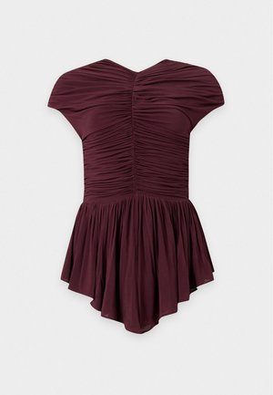 Femme debout, mains dans le dos, portant un haut froncé bordeaux foncé avec un ourlet évasé et un pantalon noir à jambes larges.
