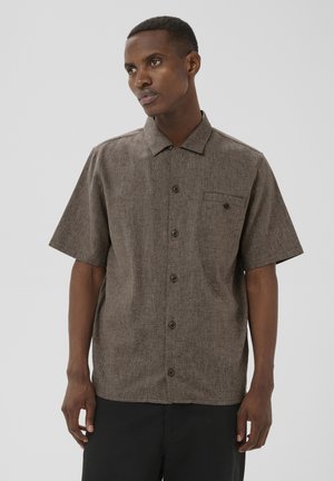 Homme portant une chemise marron à manches courtes, texturée, avec col et poche poitrine, debout devant un fond gris clair uni.