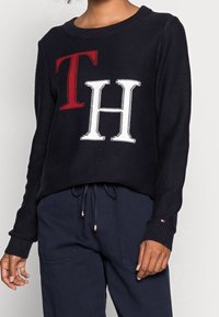 Marineblauwe gebreide trui met rode "T" en witte "H" letters op de voorkant. Brede halslijn, geribde manchetten en onderkant. Gestyled met donkere broek.