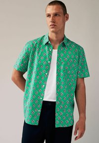 Kurzarmhemd aus leuchtend grünem Stoff mit einem sich wiederholenden Blumenmuster in Weiß und Lila, kombiniert mit einem weißen Unterhemd.