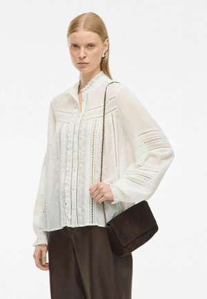 Bijela, nabraná bluzica s vertikalnim izrezima, dugim rukavima i kopčanjem na dugme, uparena s tamnosmeškom zamščenom torbom preko ramena.