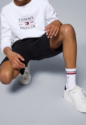 Osoba klęcząca, ubraną w biały sweter Tommy Hilfiger, czarne spodenki, białe skarpetki z czerwonymi i granatowymi paskami oraz białe sneakersy na sznurowadła.