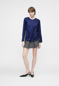 Blusa di raso navy con maniche lunghe, scollatura rotonda e dettaglio plissettato frontale, abbinata a una mini gonna plissettata grigia e scarpe con tacco slingback nere.