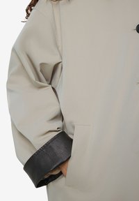 Beige jas met zwarte leren manchetten, waarbij een hand in de zijzak is gestoken, wat de textuur van de stof en de details van de knopen benadrukt.