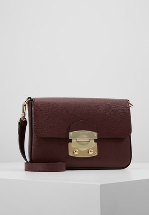 Ciemnobrązowa, teksturowana skórzana torba crossbody z metalową klamrą w złotym odcieniu i regulowanym paskiem na białym podeście na szarym tle.