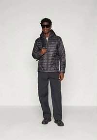 Μαύρο μπουφάν με μόνωση και φερμουάρ, διαθέτει κουκούλα και λογότυπο. Συνδυασμένο με μαύρο παντελόνι και παπούτσια, στέκεται σε απλό φόντο.