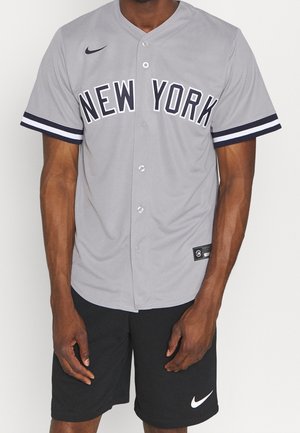 Mężczyzna w szarej koszulce baseballowej New York z guziczkami z przodu i czarnych szortach Nike z białym logo Swoosh.
