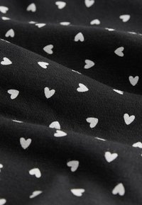 Sort stof med små hvide hjerte-mønstre, jævnt fordelt over overfladen, der viser bløde folder og tekstur.