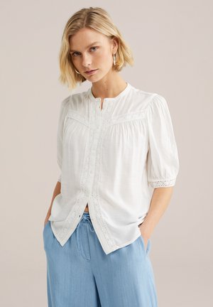 Blond žena v bielej blúzke s krátkym rukávom s čipkovými detailmi a svetlomodrých nohaviciach so šnúrkou v páse, stojaca s rukami v kapsách.