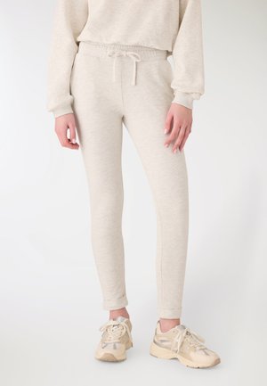 Persona che indossa jogger beige chiaro aderenti con vita regolabile a cordoncino, abbinati a sneakers beige e crema con lacci, in piedi contro uno sfondo semplice.