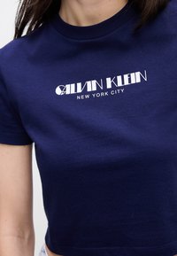 Tamsiai mėlynas medvilninis marškinėliai su trumpomis rankovėmis, su baltu grafiniu logotipu "CALVIN KLEIN NEW YORK CITY" ant krūtinės. Apvalus iškirpimas.