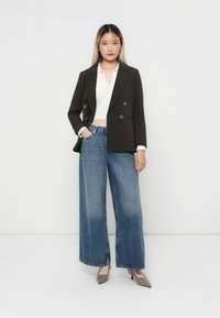 Brun dobbeltspent blazer over en hvit strikket topp, parret med høytlivte vide blå jeans og grå spisse hæler.