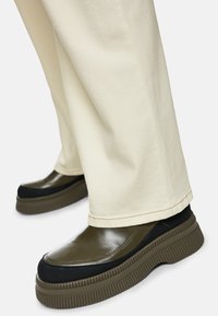 Black and olive green leather shoes with a ridged sole and smooth texture, paired with cream-colored pants, partially visible in the frame.
