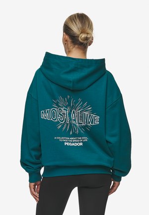 Turkos oversized hoodie med dragsko i huvan. Baksidan har grafisk text "MOST ALIVE" och sprakar. Tillverkad av mjukt tyg med ribbat avslut.