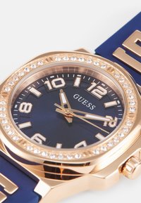 Reloj en tono dorado con una esfera azul marino, bisel adornado con cristales y marcadores de hora audaces. Presenta una correa de goma azul con acentos dorados.