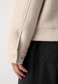 Beige texturerad jacka i ett mjukt, fleece-liknande material, med en manschett med en tryckknappdetalj, tillsammans med mörka tygerbyxor.