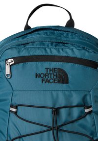 Turkusowy plecak z czarnym zamkiem, z logo The North Face i elastycznymi sznurkami bungee z przodu, wykonany z trwałego materiału.